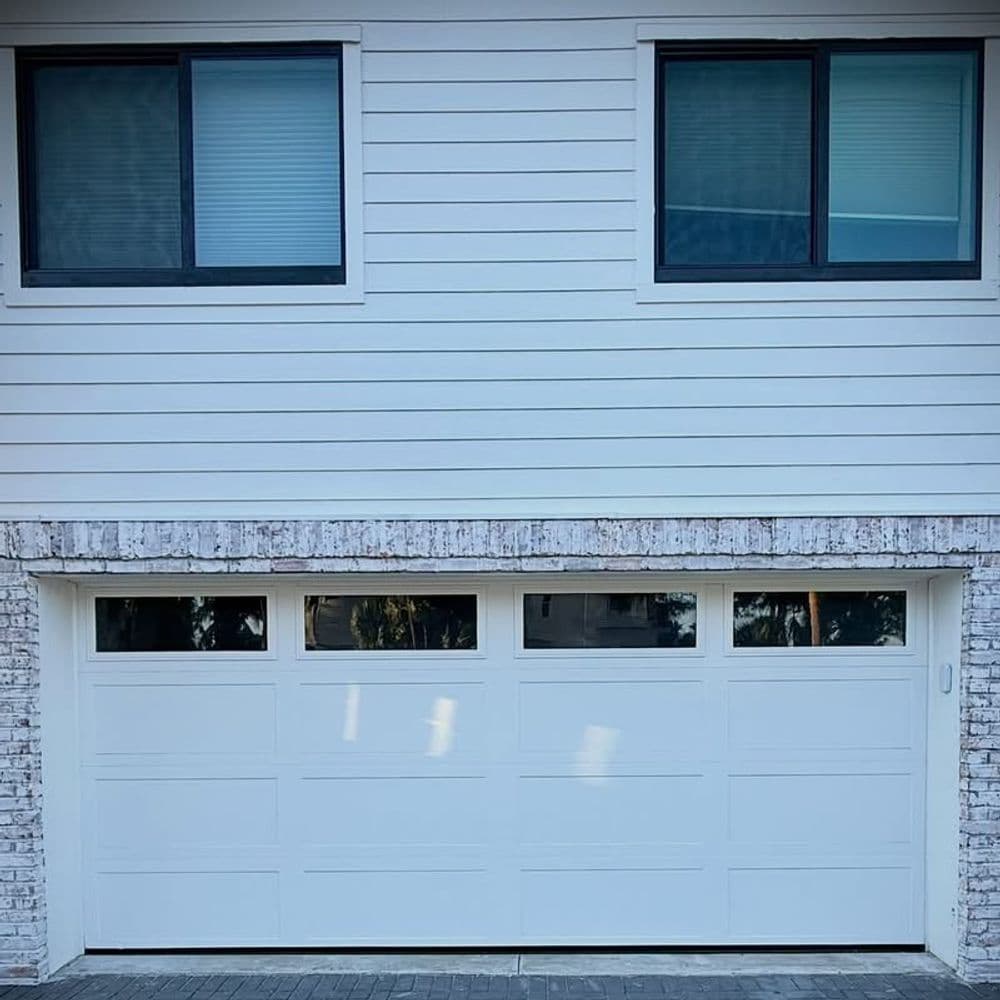 Modern white garage door with four horizontal panels, framed by brick and two windows above.