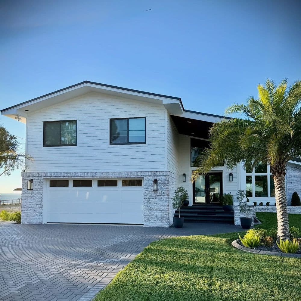 Modern white house with stone accents, palm trees, and a paved driveway on sunny day.