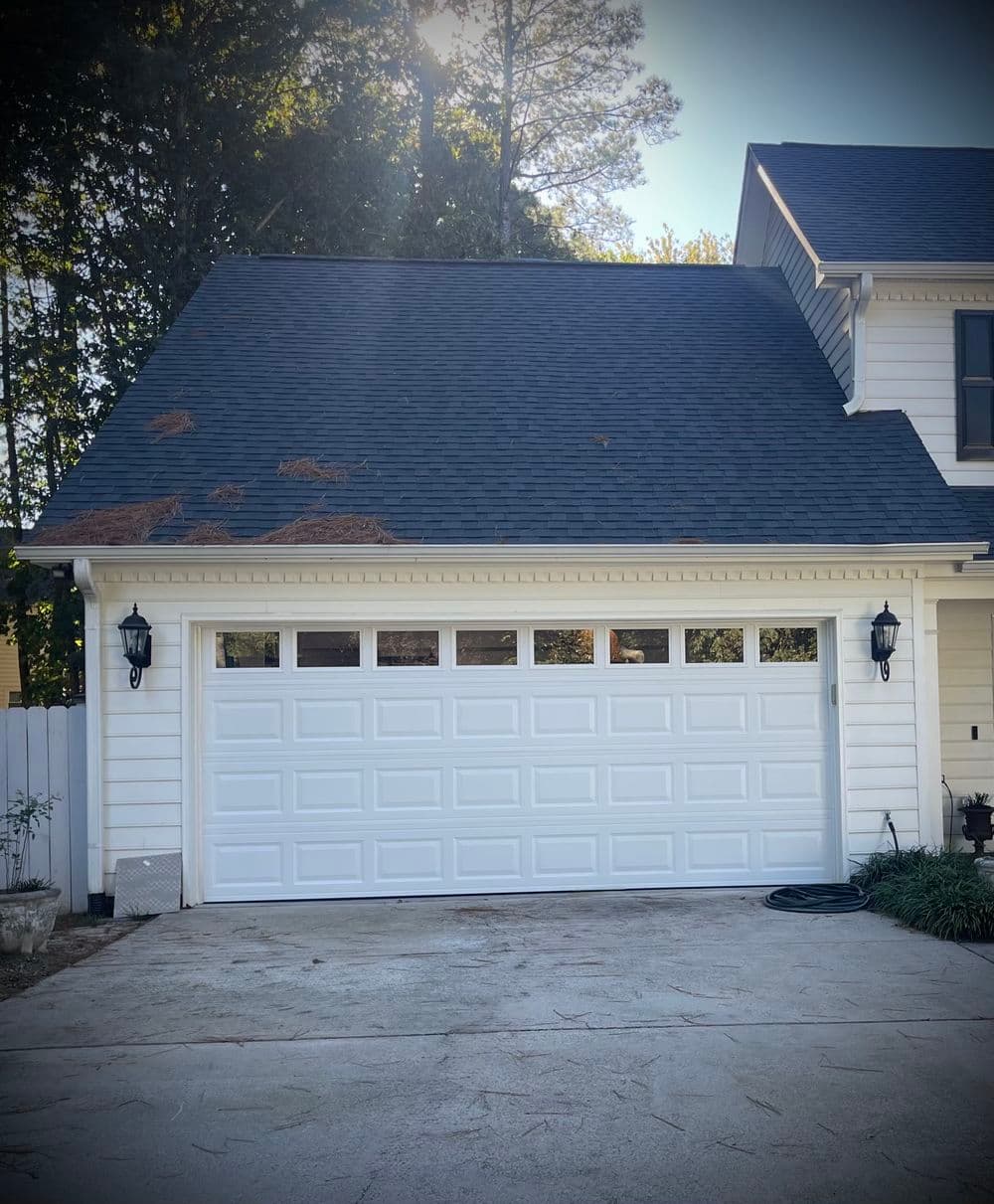 White garage door on a house with a black roof and landscaped driveway. Sunlight in background.