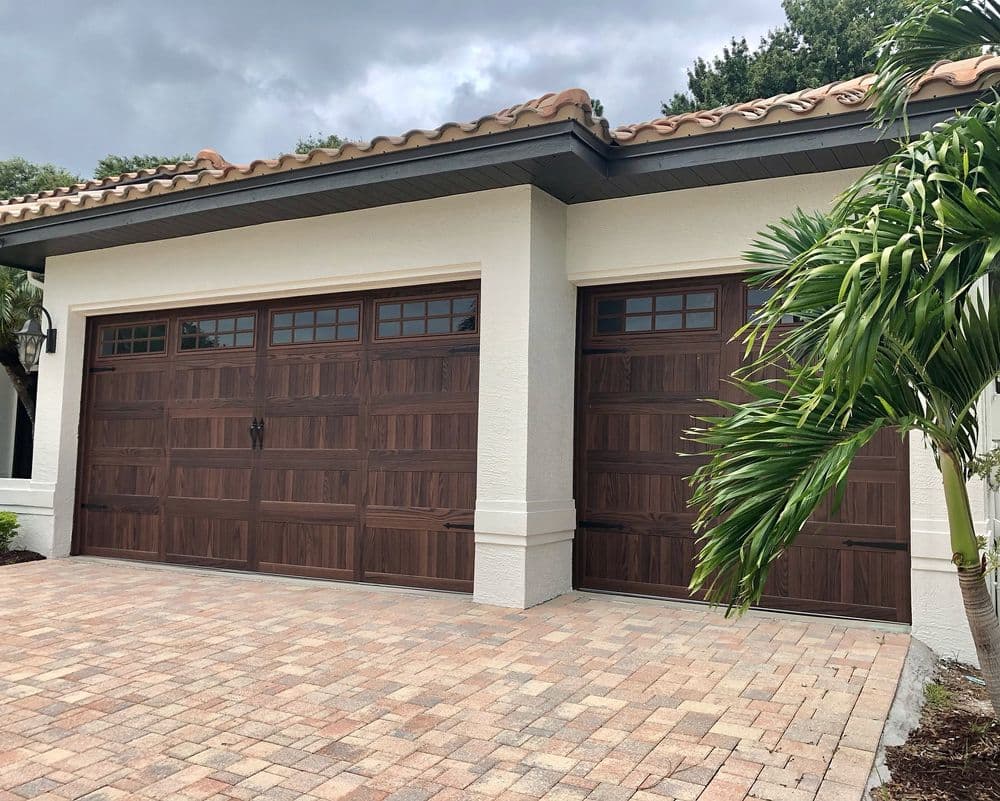 Elegant wooden garage doors on a modern home with paver driveway and tropical landscaping.