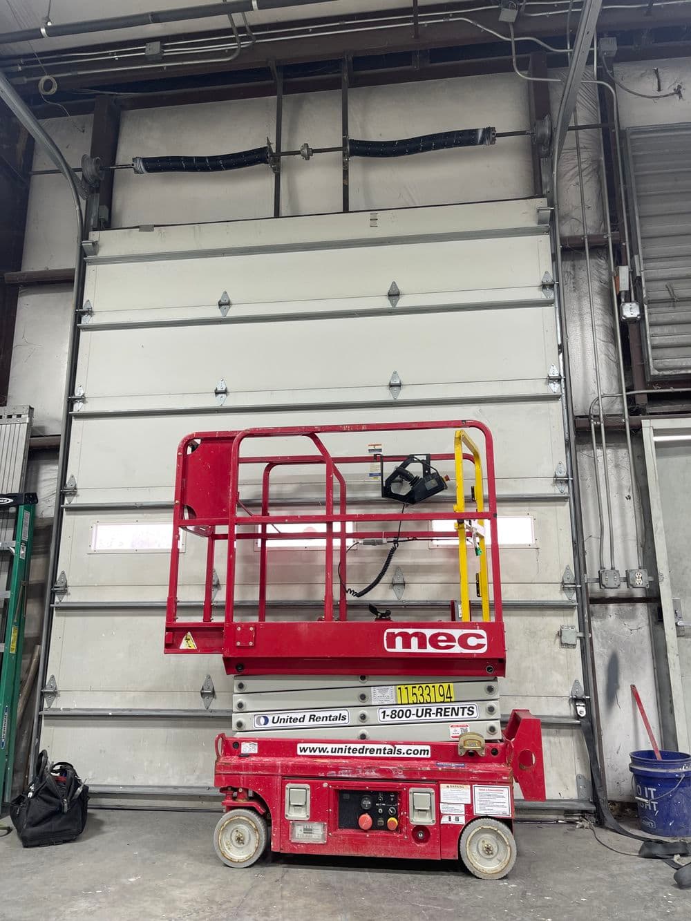 Red MEC scissor lift parked in a warehouse with a large closed garage door. United Rentals signage visible.