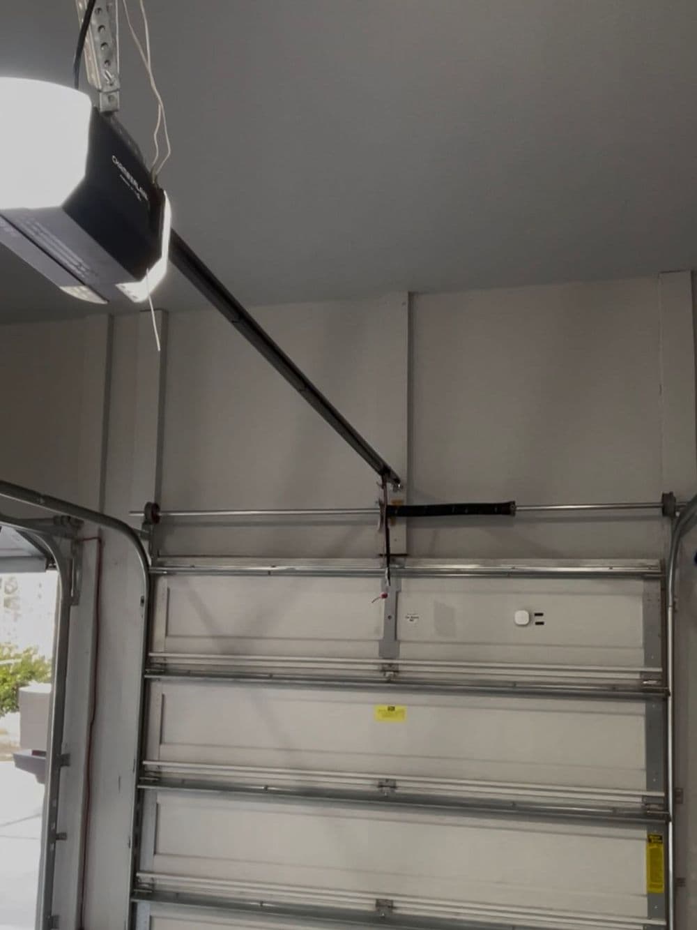 Garage door opener mounted on ceiling with rails and control panel visible, inside a garage.