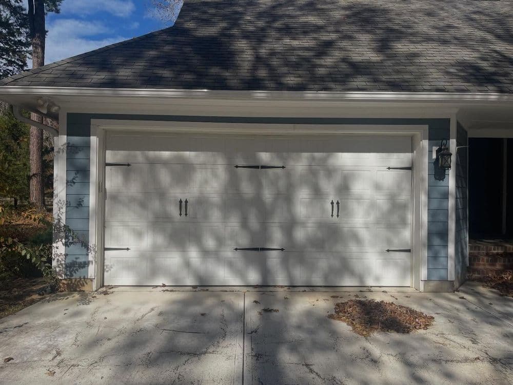 White garage door with decorative hardware, grey house exterior, and blue siding in daylight.